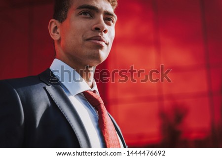 Focus on low angle of positive young businessman in tie and suit. He is standing outside and looking away thoughtfully. Copy space in right side