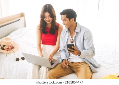 Focus On Man. Happy Smile Young Adult Southeast Asian Couple Using Laptop For Getting Ready For Holidays Travel Trip. Living At Home Sitting On Bed With Hat And Luggage.