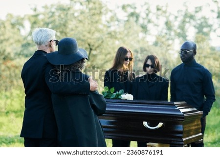 Focus on mature mourning couple wearing black attire standing in front of coffin and group of young intercultural grieving people at funeral