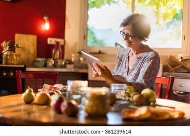 Focus On A Middle Aged Woman Looking At Recipes On A Tablet In A Red Kitchen. She Is Sitting At A Wooden Table With Fruits, Old Fashioned Jar And Utensils Around Her. Blur Background, Shot With Flare