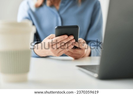 Focus on mobile phone held by young female white collar worker in blue shirt sitting by desk in front of laptop and scrolling through messages