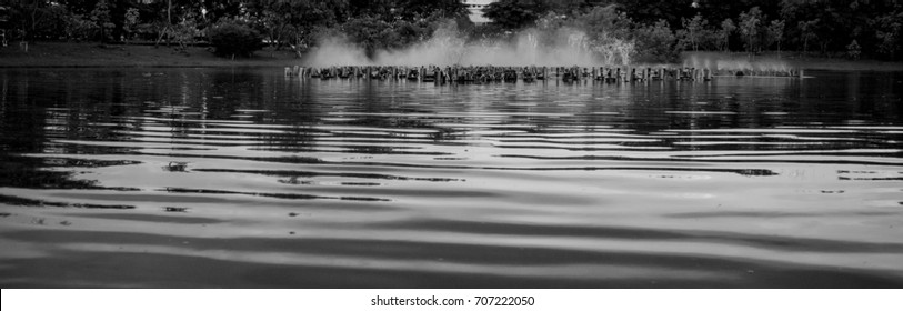Focus on patterns and textures of water ripples on the surface of a lake, with water sprays from fountain spouts in the background, in black and white.