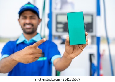 Focus On Phone, Smiling Petrol Pump Worker Showing Green Screen Mobile Phone By Pointing Finger By Looking At Camera At Filling Station - Concept Of Online Booking, Advertisement And Gasoline Servie.