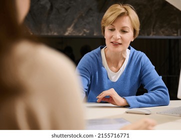 Focus On Pleasant Mature Caucasian Woman, Experienced Secretary-consultant, Advising A Client At The Front Desk Of The Registration Office Building, Medical Clinic, Sales Department Or Travel Agency