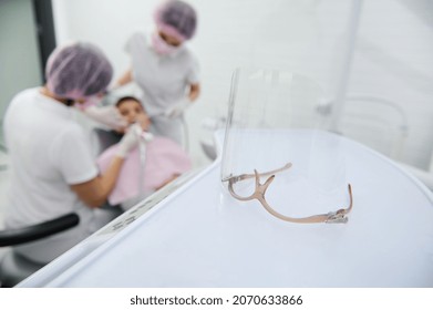 Focus On Safety Goggles, Protective Medical Transparent Mask For Health Care Worker Used In Dentistry Against Blurred Background Of Pediatric Dentist And Assistant Treating Child's Oral Cavity