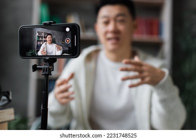 Focus on screen of smart phone, asian man in casual clothes talking and gesturing while recording video on modern phone on tripod. Male blogger sitting at home and doing live stream. - Shutterstock ID 2231748525