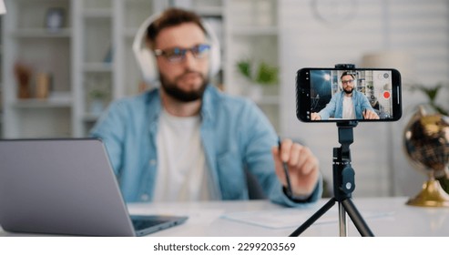 Focus on screen of smartphone. Young man is live streaming a training blog at home. Blogger using a smartphone while sitting at home in the kitchen with a laptop. - Powered by Shutterstock