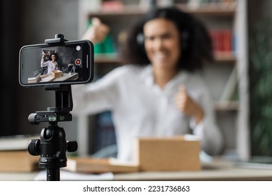 Focus on screen, young multinational woman recording video on phone camera while unpacking box with new wireless headphones showing thumbs up. - Powered by Shutterstock