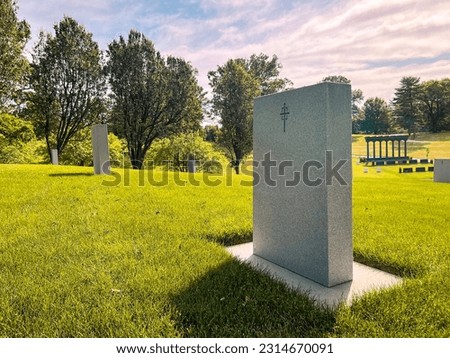 Focus on a single empty tombstone in a row of unmarked gravesites. The front of the stone is blank, except for a small engraved cross at the top of the headstone. Situated in a lush green cemetery. 