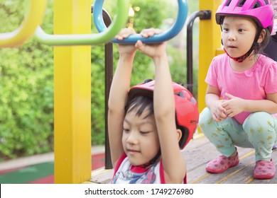 Focus On Sister. Two Cute Little Asian Sibling Hanging On The Monkey Bar In Playground. Two Asia Kid Wearing Helmet To Protect His Head. Concept Playing Outdoor, Adventure Activity, Safety And Fun.