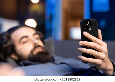 Focus on smartphone with man in blurry background lounged on couch texting friend inviting him to watch movie on streaming service. Close up on phone used by person at home discussing with mate - Powered by Shutterstock