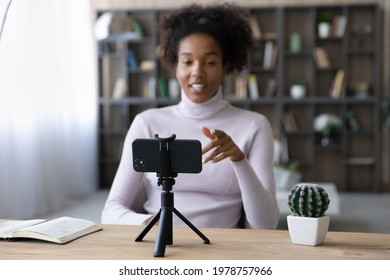 Focus on smartphone standing on table on tripod stabilizer, recording african american female blogger. Smiling pretty young mixed race woman influencer filming educational video on phone web camera.