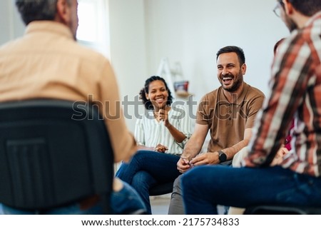 Focus on the smiling man, talking with people of all ages, during the group therapy.