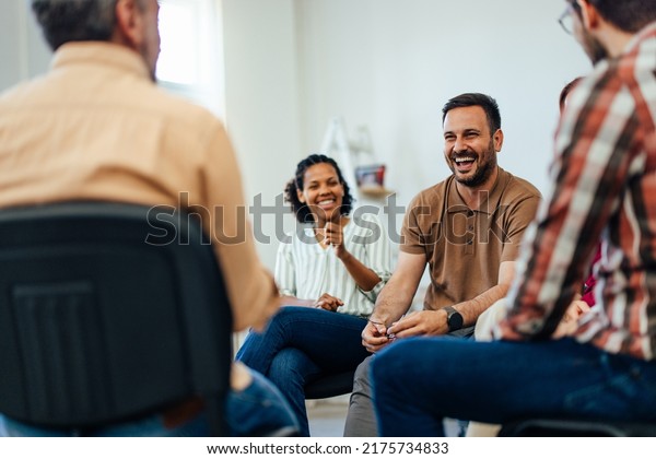 Focus on the smiling man, talking with\
people of all ages, during the group\
therapy.