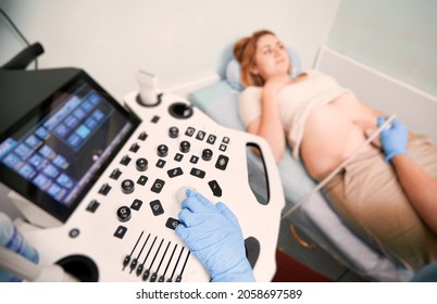 Focus on sonographer's hand in sterile gloves performing ultrasound scanning in clinic. Woman lying on daybed while doctor examining patient abdomen with ultrasound scanner on blurred background.