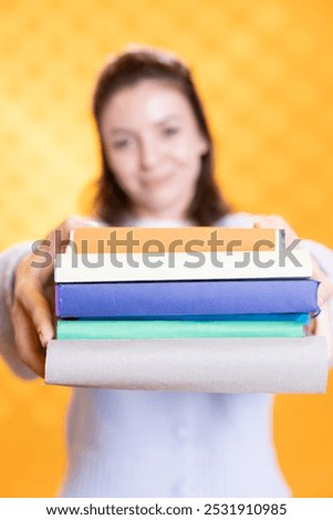 Focus on stack of books held by woman in blurry background doing recommendation, isolated over studio background. Close up shot of pile of novels offered by cheerful nerd doing endorsement