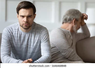 Focus on stressed angry young man sitting separate on couch with stressed offended middle aged father. Unhappy grownup son feeling irritated after argue quarrel with dad, generations gap concept.