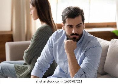 Focus on stressed thoughtful young man sitting separate from offended wife on couch at home. Young frustrated married family couple ignoring each other after quarrel, relations problems concept.