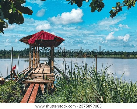 focus on the structure of the red-roofed wooden jetty above the river. there are green leaves above and grass below. the background is the river, distant trees and a cloudy blue sky
