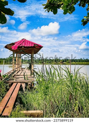 focus on the structure of the red-roofed wooden jetty above the river. there are green leaves above and grass below. the background is the river, distant trees and a cloudy blue sky