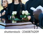 Focus on two fresh white roses held by mourning man in black suit during funeral service while standing by coffin against his daughter and wife