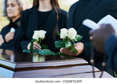 Focus on two fresh white roses held by mourning man in black suit during funeral service while standing by coffin against his daughter and wife