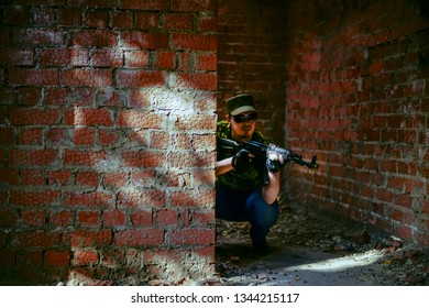 Focus On Wall. Caucasian Military Man With Black Sunglasses Indoor Urban Room Space Stand With Machine Gun Near Abandoned Red Brick Wall.Dusk Light. Corridor In Perspective.Empty Space For Inscription