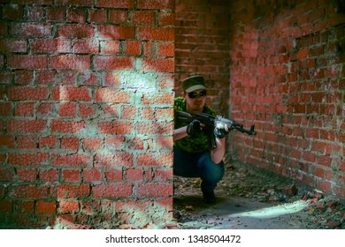 Focus On Wall. Caucasian Military Man With Black Sunglasses Indoor Urban Room Space Stand With Machine Gun Near Abandoned Red Brick Wall.Dusk Light. Corridor In Perspective.Empty Space For Inscription