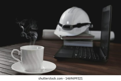Focus On White Ceramic Coffee Cup With Steam And Blurred Background Of Black Laptop And White Safety Helmet On Engineer Office Desk In Vintage Tone Style