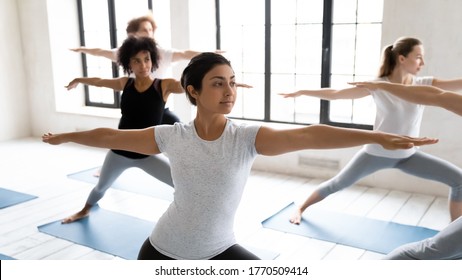 Focus On Young Fit Indian Ethnicity Woman Standing In Virabhadrasana Second Position, Practicing Warrior II Exercise With Diverse Motivated People At Group Yoga Master Class In Modern Studio.