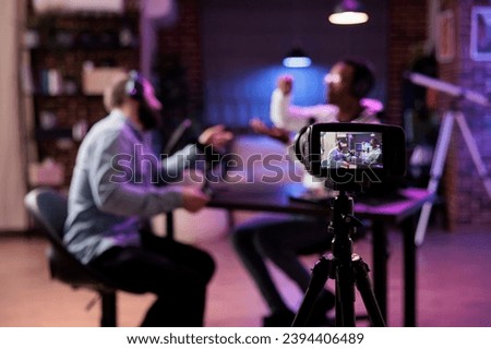 Focus shot on professional camera used to record podcast show with presenter and guest discussing in blurry background. Equipment in home studio used for live streaming broadcast