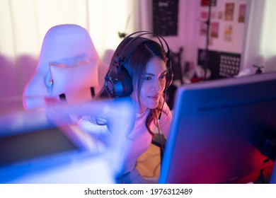 Focus of smiling young pretty Asian transsexual transgender professional gamer wearing a headset sitting on a gaming chair playing online video games on a personal computer in a violet neon room. - Powered by Shutterstock
