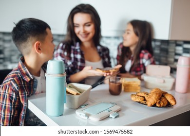 Focus At Thermos On Table, Behind Young Mother And Her Kid Making School Sandwich For Lunch.