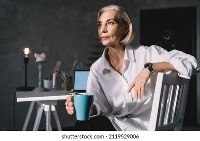 Focused 50 Years Old Caucasian Businesswoman With Cup Of Coffee Sitting On Chair And Looking Away. Small Business And Entrepreneurship. Modern Successful Woman. Dark Art Studio With Sunny Lighting