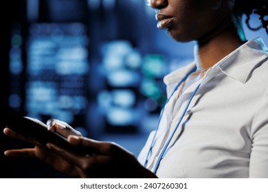 Focused admin in data center uses tablet to prevent server machines overload. Expert between server mainframes using device to ensure enough network bandwidth for smooth operations