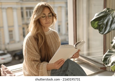 Focused adult caucasian woman reading book at home sitting by window in warm weather. Pretty woman wears sweater glasses and jeans. Leisure, literature and people concept