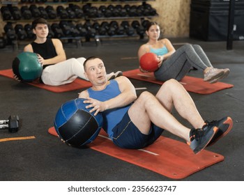 Focused adult man taking part in high-intensity group training session, doing V-sit exercise with medicine ball and twisting torso to strengthen abdominal and oblique muscles.. - Powered by Shutterstock