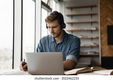 Focused Adult Student Guy In Headphones Listening To Online Learning Seminar, Preparing For Virtual Exam, Passing Language Course, Watching Webinar On Laptop, Taking Notes, Studying At Home