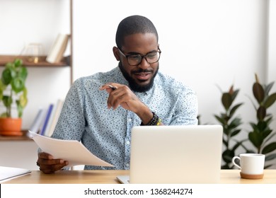 Gerichte Afro-Amerikaanse zakenman die werkt met laptopdocumenten in kantoor met papieren die rapporten voorbereiden die werkresultaten analyseren, zwarte mannelijke analist die papierwerk doet op de werkplek met behulp van computer