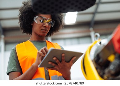 Focused african american female engineer wearing safety goggles and using digital tablet while controlling robotic arm in factory