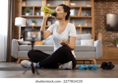 Focused african american female in gym clothes drinking water from sports bottle while sitting cross-legged in front of active cell phone on tripod. Fit sports vlogger showing workout routine at home. - Powered by Shutterstock