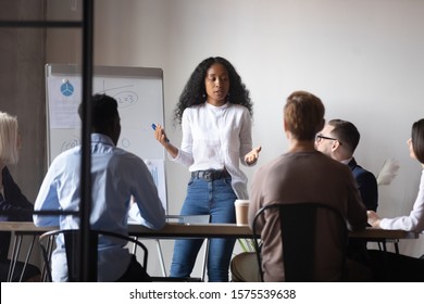Focused african american female speaker presenting project research results to interested diverse teammates at brainstorming meeting. Confident mixed race coach giving educational lecture or workshop. - Shutterstock ID 1575539638
