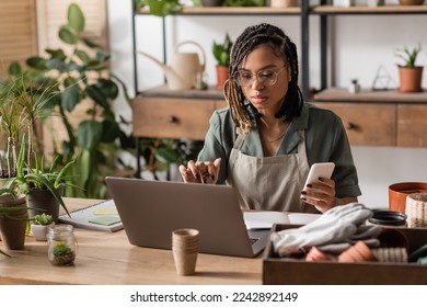 focused african american florist sitting at workplace and smartphone   notebook in flower shop