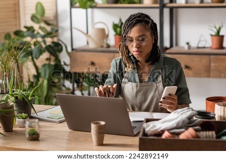 focused african american florist sitting at workplace with smartphone and notebook in flower shop