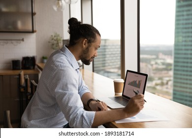 Focused African American Male Employee Sit At Desk In Office Work On Computer Analyzing Graphs. Concentrated Biracial Man Worker Look At Laptop Screen Busy With Paperwork At Workplace.