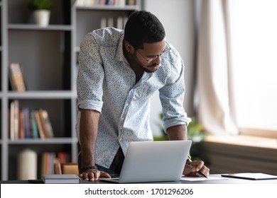 Focused African American Male Worker In Glasses Stand At Office Desk Busy Working On Laptop Gadget, Concentrated Biracial Man Make Notes Planning Considering Business Project On Computer Device