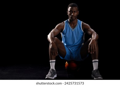 Focused African American man poses on a basketball, indoor setting on a black background with copy space. His intense gaze and athletic posture convey determination and strength. - Powered by Shutterstock