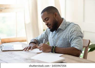 Focused African American Man Student Freelancer Making Notes Studying Working With Laptop, Young Black Man Professional Writing Essay In Notebook Preparing For Test Exam Sit At Home Office Desk