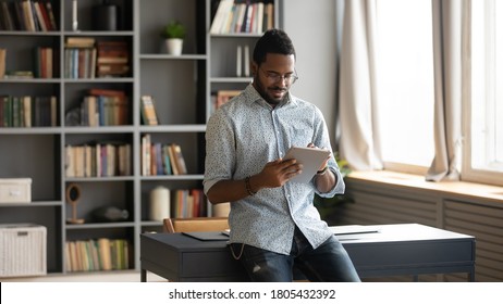 Focused African American man using computer tablet, standing in modern cabinet new work desk, looking at screen, confident young male wearing glasses checking email, working online, writing message - Powered by Shutterstock