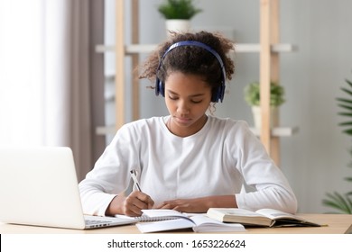 Focused african american teenage girl wearing headphones writing notes study with laptop and books, serious black female high school teen student listening audio course or music while doing homework